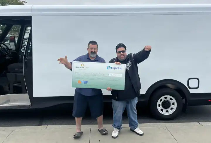 Two fleet owners holding an oversized $96,750 HVIP voucher check beside a BrightDrop electric van